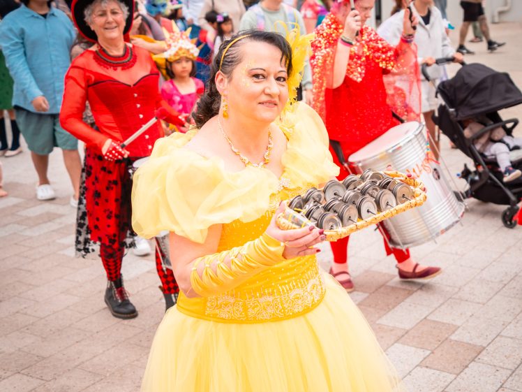 Image of A woman in a yellow princess dress holds a tray of cupcakes and leads a colourful parade with others in bright costumes, including red outfits and hats, as people watch and children ride in a pushchair.