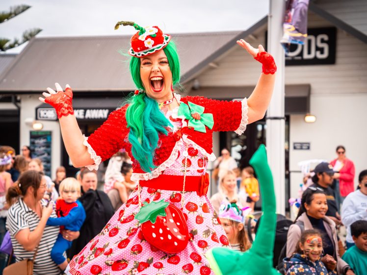 Image of A smiling performer in a strawberry-themed outfit with a green wig entertains a crowd at an outdoor event. People, including children, watch and smile in the background.