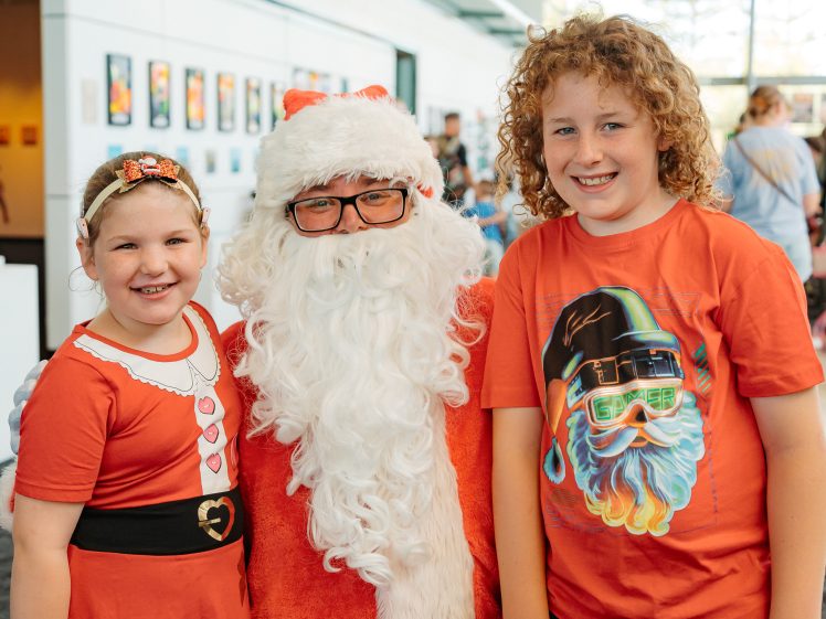 Image of A person dressed as Father Christmas poses with two smiling children. The girl on the left wears a red dress and a headband, while the boy on the right wears a red shirt with a colourful Father Christmas graphic. They are indoors with art on the wall behind them.