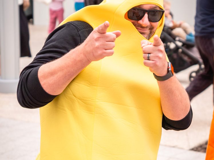 Image of A person wearing sunglasses and a bright yellow banana costume points playfully at the camera, surrounded by people in an outdoor setting.