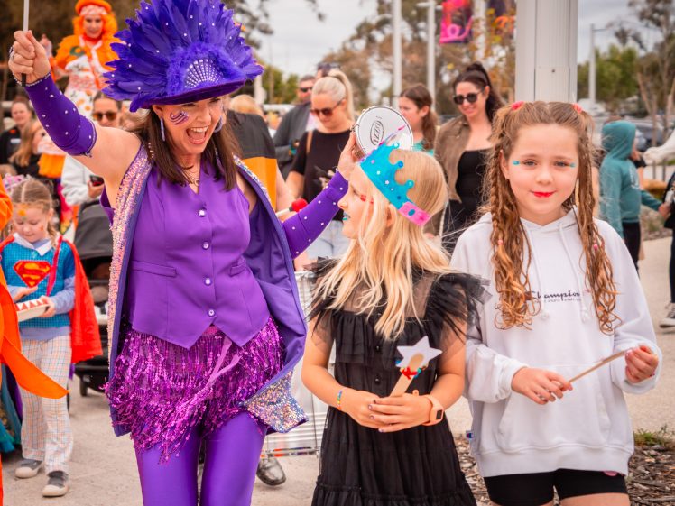 Image of A joyful parade scene with a woman in a vibrant purple costume dancing alongside two young girls, one in a black dress and one in a white hoodie. A crowd, some in colourful costumes, follows in the background.