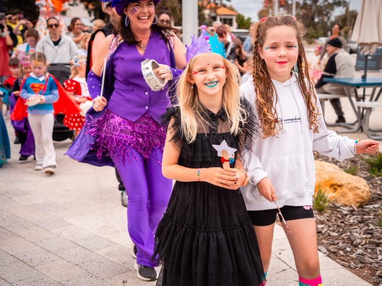Image of A group of children and adults in colourful, festive costumes parade outdoors. Two girls lead, one in a black dress with a blue crown and wand, the other in white with rainbow socks. An adult in purple follows, smiling and holding a tambourine.