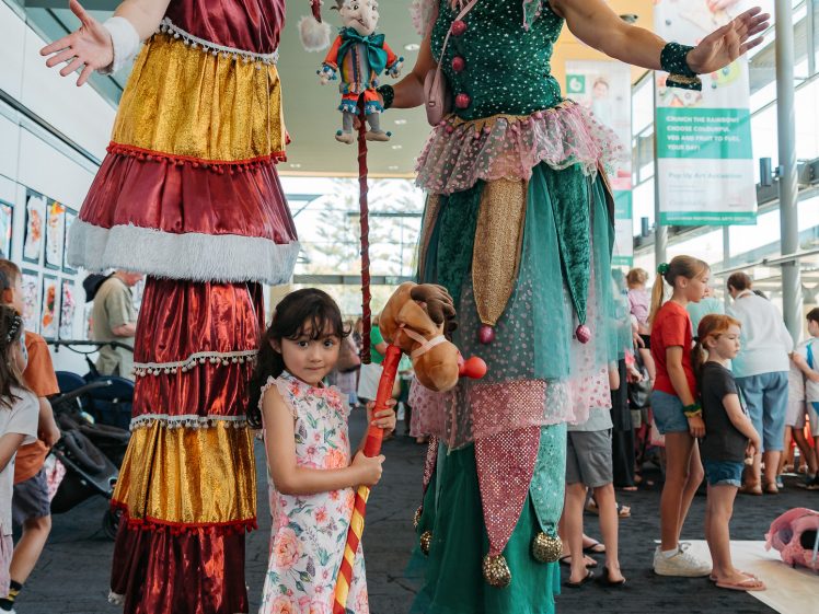 Image of Two women in colourful jester costumes stand on stilts, smiling and posing with a young girl holding a toy horse. The scene is indoors with people and artwork in the background.