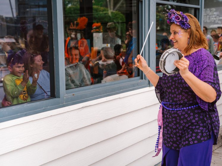 Image of A woman dressed in purple with a flower crown plays a tambourine outside a window while smiling children and adults watch and cheer from inside. Bright costumes and happy faces are visible through the glass.