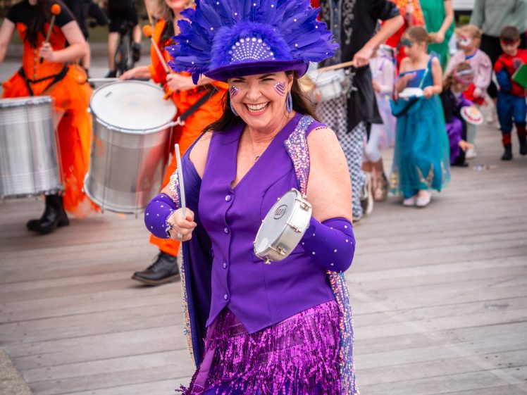 Image of A woman in a bright purple costume and feathered headdress smiles while playing a tambourine in a festive parade, with other colourfully dressed performers and children in fancy dress in the background.