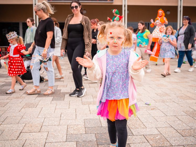 Image of A young girl in colourful clothes and face paint stands clapping in the foreground, whilst adults and children in fancy dress walk behind her near shopfronts and a café.