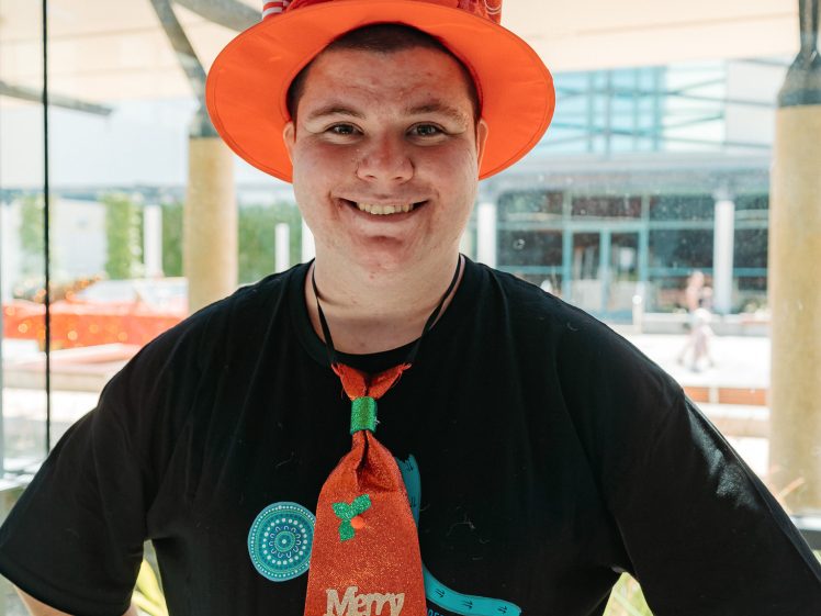 Image of A smiling person stands indoors, wearing a bright red Christmas hat with green decorations and a red Merry Christmas tie, over a black T-shirt with blue designs. Sunlight streams in through large glass windows behind them.
