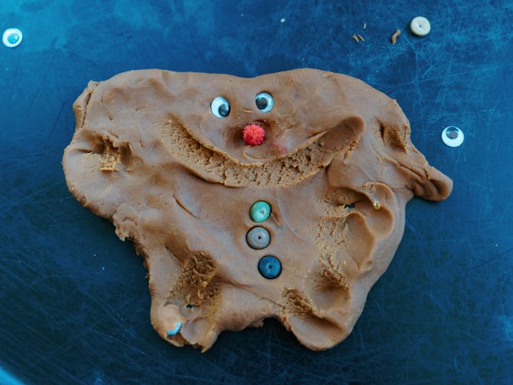 Image of A misshapen, playful brown playdough figure with a big smile, button eyes, a red pom-pom nose, and three buttons down its front, on a scratched blue surface.
