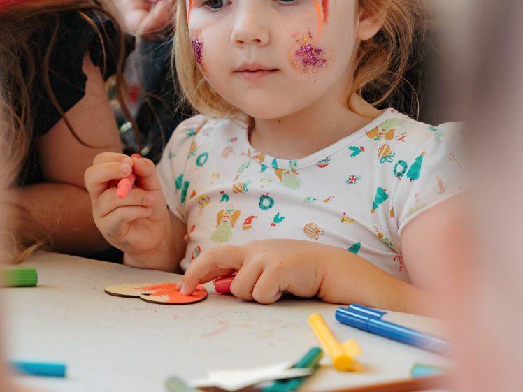 Image of A young child with colourful face paint and a glitter sticker is sitting at a table covered with felt-tip pens and paper, appearing focused whilst holding a red felt-tip pen and participating in a creative activity.