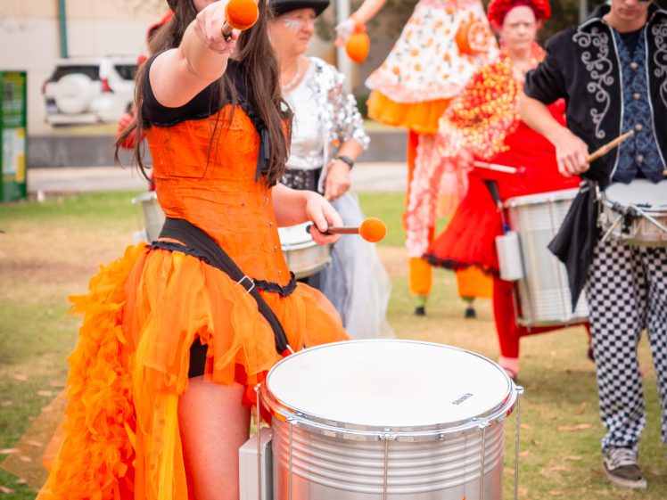 Image of A woman in an orange costume plays a large drum outdoors, surrounded by other performers in colourful outfits, including a stilt walker in a matching dress. Trees and parked cars are visible in the background.