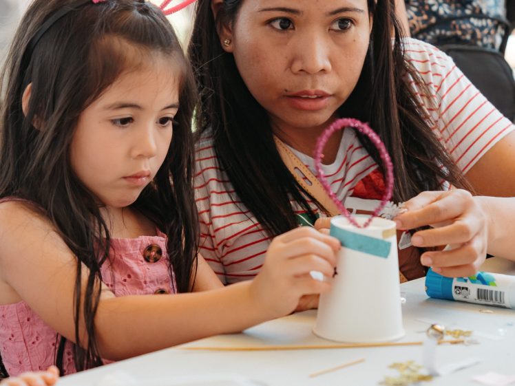 Image of A woman and a young girl sit at a table, focused on making a craft project together. The girl wears pink and has headband accessories; the woman wears a red headband. Art supplies are scattered on the table.