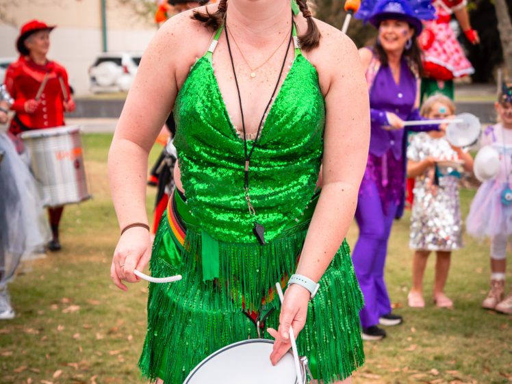Image of A woman in a sparkly green outfit and top hat plays a drum outdoors, smiling. Behind her, people in colourful costumes are also celebrating, suggesting a festive parade or event.