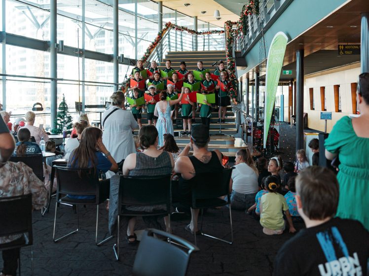 Image of A group of people dressed as elves sing on a staircase decorated with Christmas garlands, while an audience sits and watches in a spacious, modern room with large windows.