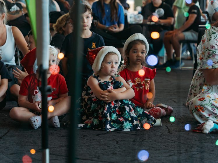 Image of A group of young children, some wearing Father Christmas hats, sit on the floor among adults. The children appear to be watching something intently. Colourful holiday lights are out of focus in the foreground.