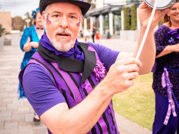 Image of A man dressed in vibrant purple clothes, with face paint and a top hat, plays a handheld drum outdoors. He is smiling and looking at the camera, with people and a building visible in the background.