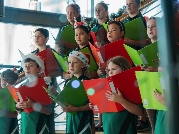 Image of A group of children in green uniforms sing together on stairs, holding red and green folders. Some wear festive headbands or hats, suggesting a holiday performance in a brightly lit indoor space.