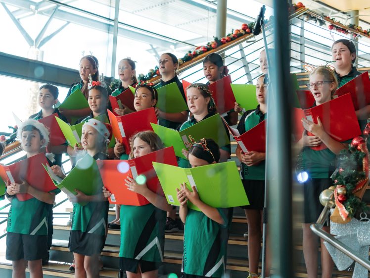 Image of A group of children in green uniforms stand on stairs, holding colourful folders and singing. Holiday decorations with garlands and ornaments line the handrails, suggesting a festive performance.