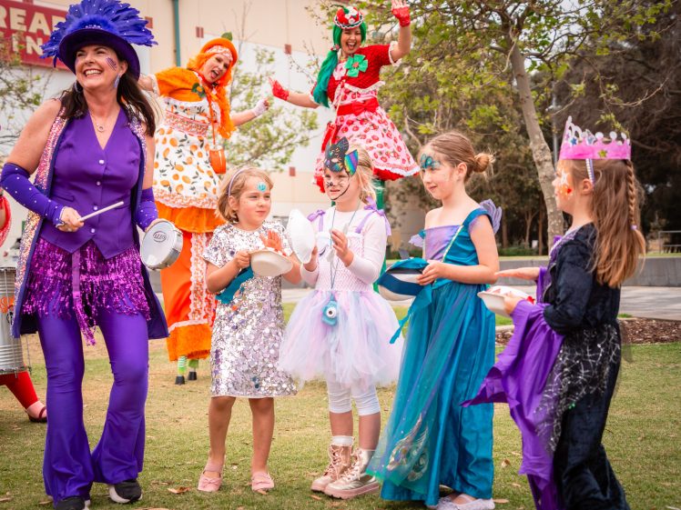 Image of A group of children in colourful costumes stand outdoors with a woman in a purple outfit, while two adults on stilts dressed as clowns entertain in the background. The scene is festive, playful, and joyful.