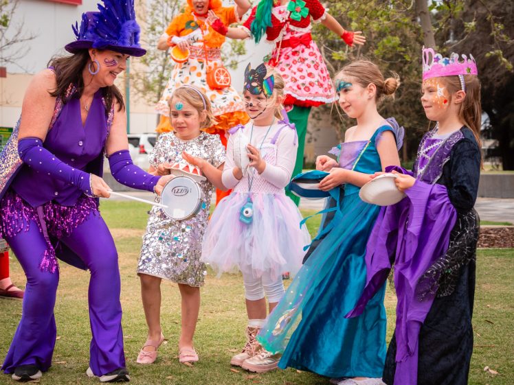 Image of A group of children in colourful costumes stand on grass with a woman dressed in a purple outfit. Two adults in bright costumes cheer in the background near a cinema building. The children appear to be playing or participating in an activity.