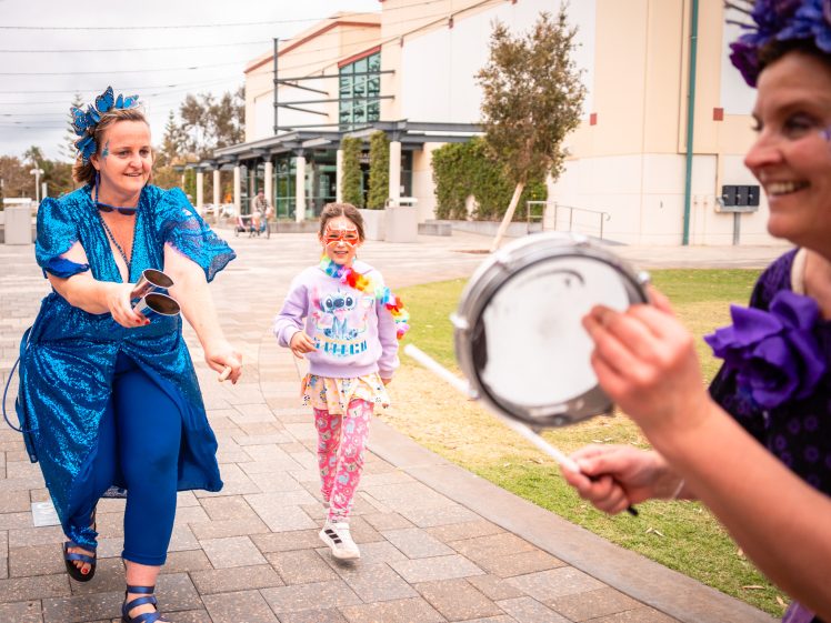 Image of Two women in blue costumes and a young girl with face paint are playing music and dancing outdoors near a modern building, creating a festive, joyful atmosphere.