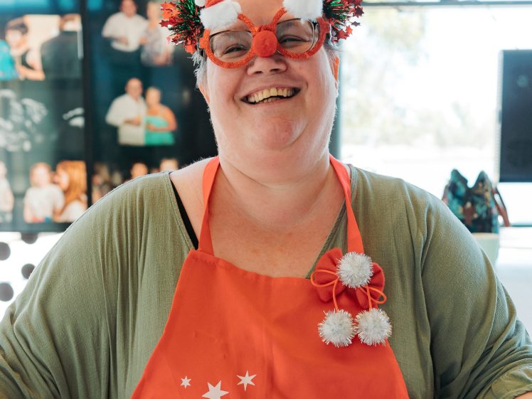 Image of A smiling person wearing festive glasses with pom-poms, a red apron that says merry, and a green jumper, standing indoors with photos and sunlight in the background.