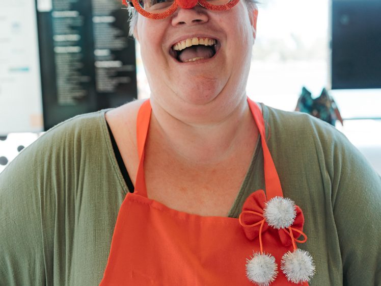 Image of A smiling person wearing festive glasses with Christmas decorations and a red apron that says merry, standing indoors with bright light in the background.