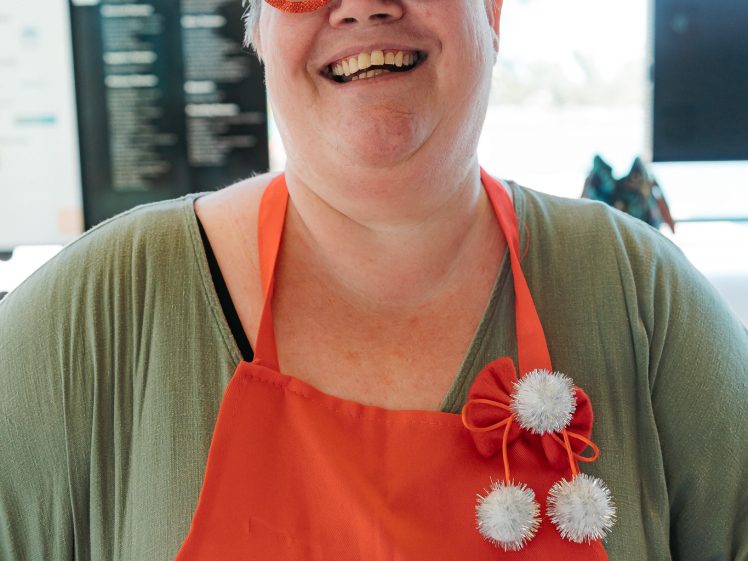 Image of A smiling person wearing festive Christmas glasses and a red apron that says “merry” stands indoors. The glasses have decorations like Santa hats and pom-poms. The person appears cheerful and joyful.