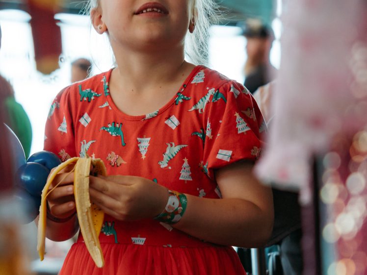 Image of A young girl in a red dress with colourful animal prints holds a peeled banana and looks upward with a smile, standing indoors with blurred people and objects in the background.