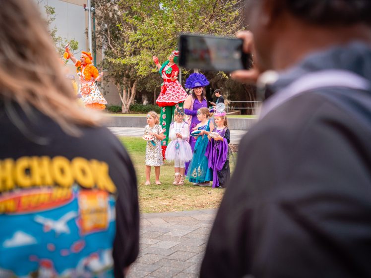 Image of Children in colourful costumes stand on grass, posed for a photo with two tall performers on stilts in bright outfits. Two adults in the foreground take pictures, one wearing a shirt with a colourful cartoon design.