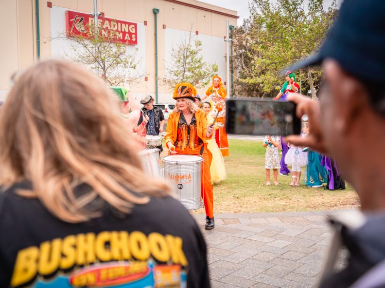 Image of A lively street performer in an orange costume plays drums for a crowd outdoors; people watch, some taking photos, and a building with a Reading Cinemas sign is in the background.