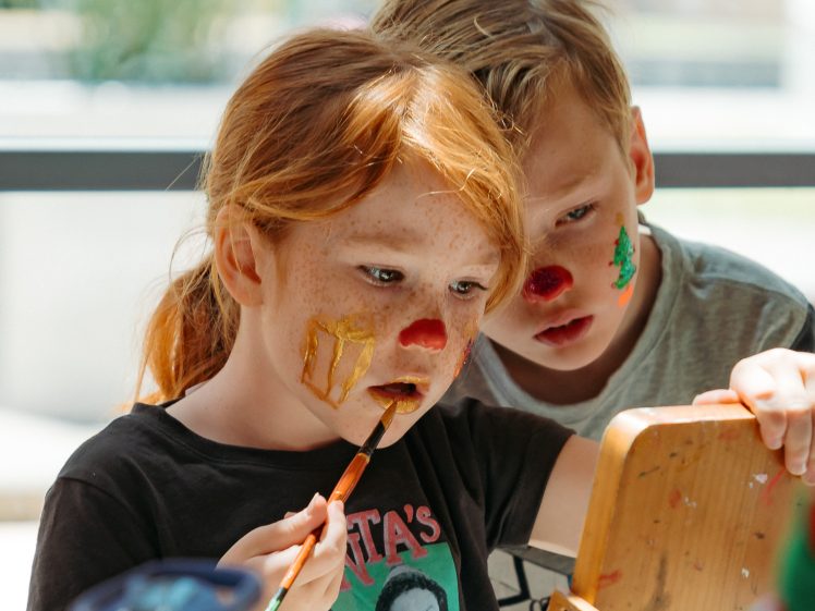 Image of Two children with face paint look at a mirror. The girl, with red hair and freckles, paints her cheek while the boy, with blond hair, watches closely. Both have red dots on their noses and colourful designs on their faces.