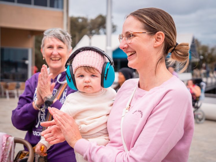 Image of A smiling woman holds a baby wearing blue noise-cancelling headphones and a pink hat. An older woman claps beside them. They are outdoors on a cloudy day, with buildings in the background.