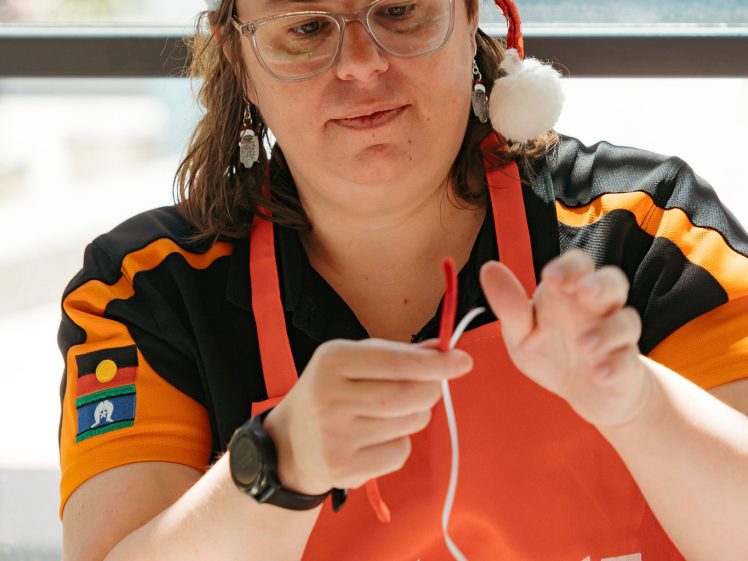 Image of A person wearing a Santa hat, red apron, and glasses is seated indoors, holding and twisting a red and white string. Festive decorations and sunlight are visible in the background.