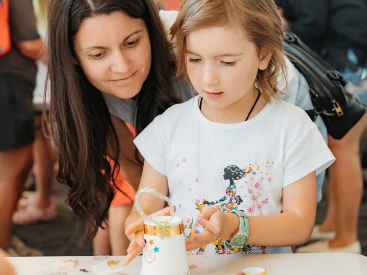 Image of A young girl decorates a white mug with gold and star-shaped stickers at a craft table, while an adult woman leans in closely to assist her. Other people are gathered and working on crafts in the background.