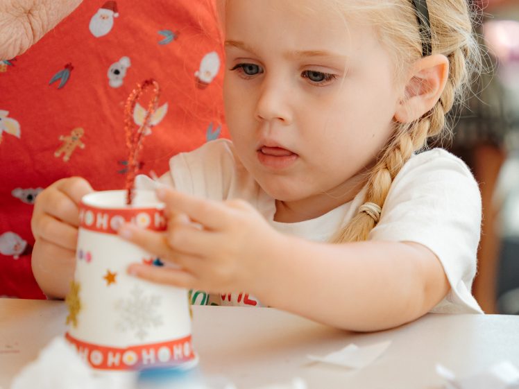 Image of A young girl with blonde plaits, wearing a reindeer antler headband, focuses on decorating a white cup with red “HO HO HO” text. She sits at a table with craft supplies, and a person in festive clothing is beside her.