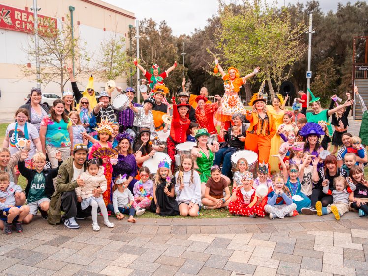 Image of A large group of adults and children in colourful costumes gather outdoors, smiling and posing for a group photo at what appears to be a festive event or celebration. Trees and buildings are visible in the background.