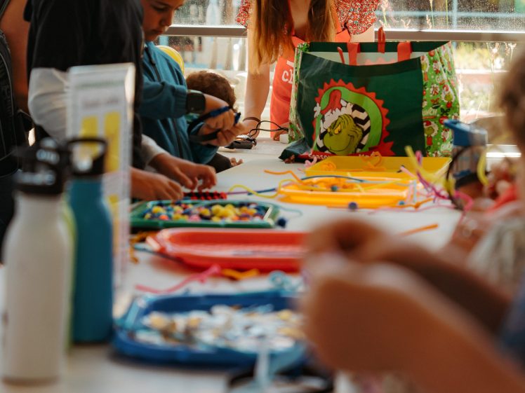Image of Children and an adult gather around a table covered with colourful craft supplies, including pipe cleaners and beads, engaging in arts and crafts activities indoors with sunlight streaming through large windows.