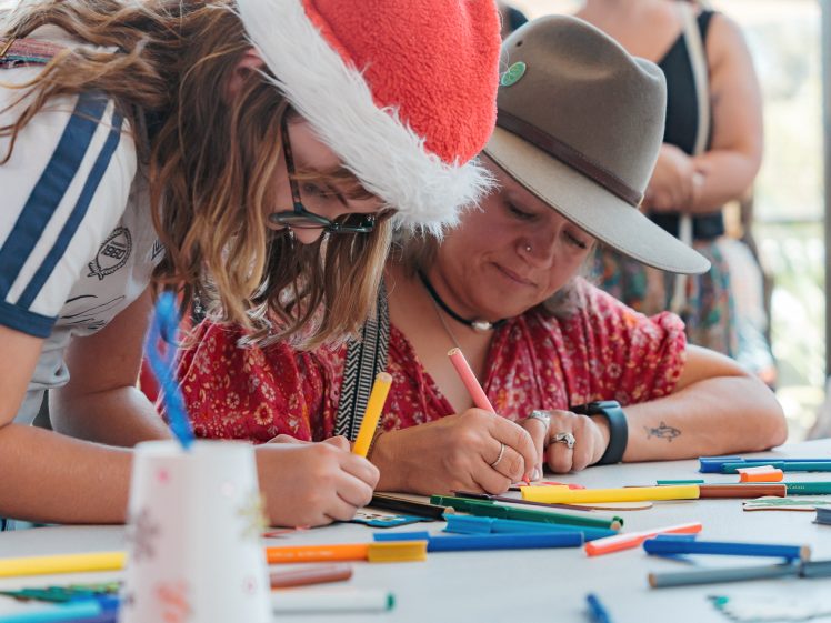 Image of Two people sit at a table drawing with coloured markers. One wears a Santa hat and glasses, and the other a wide-brimmed hat and red patterned shirt. Art supplies are scattered on the table, and others are in the background.