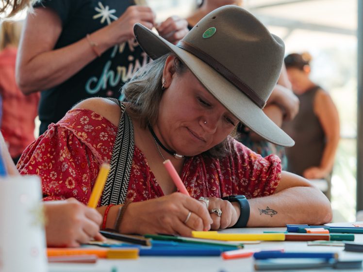 Image of A woman in a wide-brimmed hat and red patterned shirt focuses on drawing with markers at a table covered in colourful pens, with other people blurred in the background.