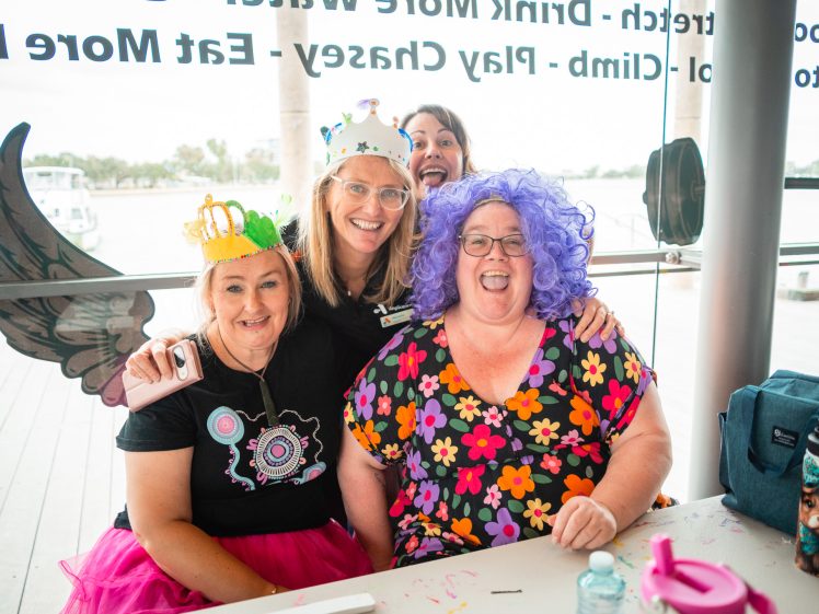 Image of Three women in colourful costumes and fun headpieces sit at a table, smiling, with one woman in a purple wig and floral dress. Another woman stands behind them, also smiling. The background shows water and large windows.