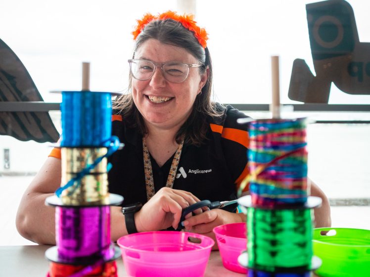 Image of A smiling woman with an orange headband sits behind a table with colourful spools of ribbon and pink bowls. She is inside near a window with event text visible in the background.