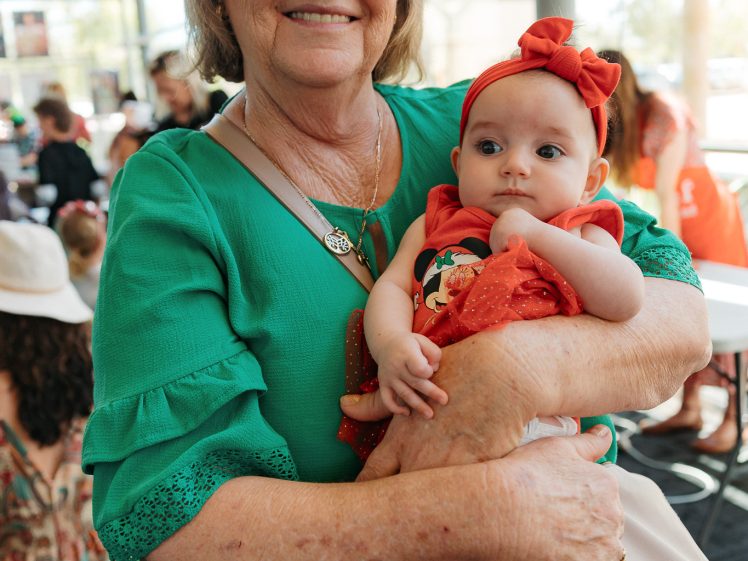 Image of An older woman smiling and holding a baby who is wearing a red hairband and dress. They are indoors in a bright, busy area with people sitting and talking in the background.