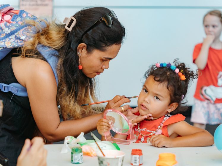 Image of A woman paints a young girl's face at a table. The girl wears a red dress and colourful hair ties, holding a mirror whilst looking at her reflection. Other children are visible in the background. Arts and crafts supplies are on the table.