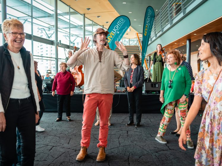 Image of A group of adults happily dancing indoors near a stage, with one man in a tan hat and red trousers leading the activity. People are smiling and enjoying themselves in a bright, modern venue.