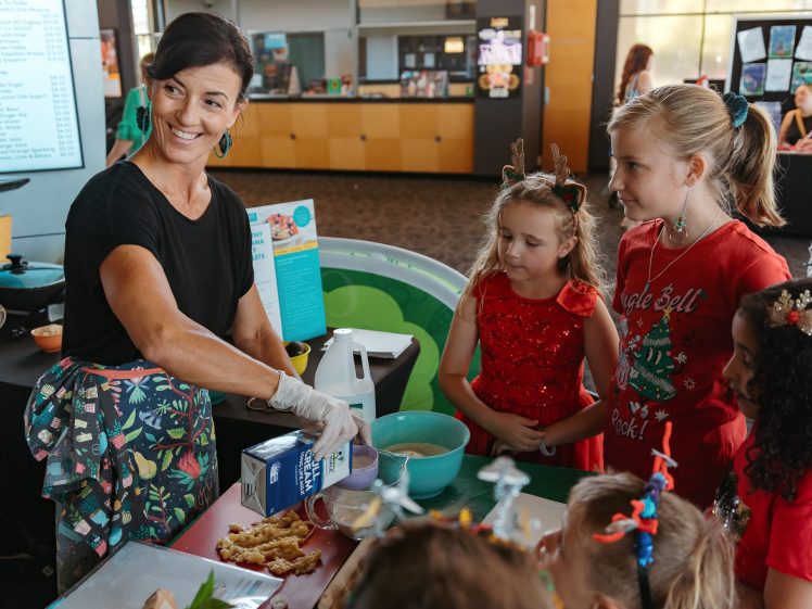 Image of A woman in an apron pours milk from a carton whilst smiling at a group of children dressed in festive, holiday-themed outfits standing around a table with baking supplies.