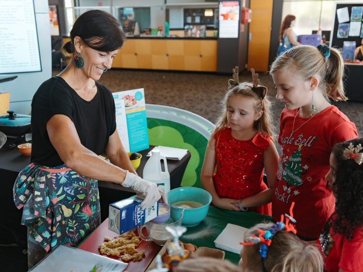 Image of A woman wearing gloves pours milk into a bowl while several children, some dressed festively, watch her at a table covered with baking ingredients in a bright indoor setting.