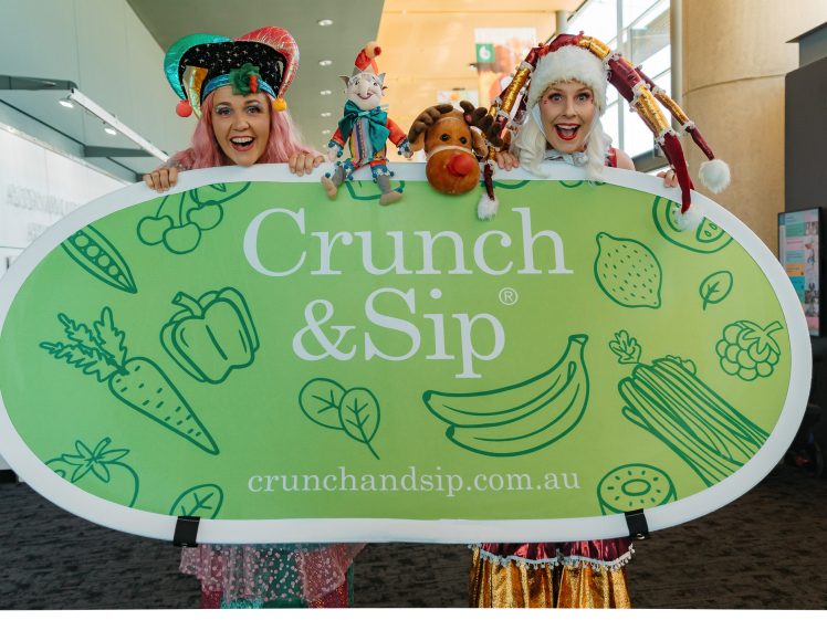 Image of Two people in colourful jester costumes smile and hold a large green Crunch & Sip sign decorated with fruit and vegetable illustrations. They have playful puppets in their hands and stand in a bright indoor space.