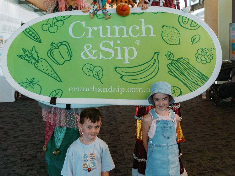 Image of Two colourful performers on stilts stand behind a large green Crunch & Sip sign with fruit and vegetable illustrations. In front, a smiling boy and girl pose together inside a bright, indoor setting.