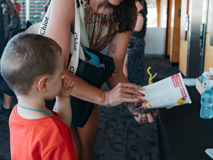 Image of A woman with glasses and a decorative headband shows a booklet to a young boy in a red shirt indoors. The woman is smiling and the boy looks at the booklet, both standing near a black table.