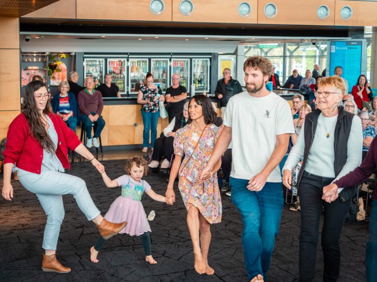 Image of A group of four adults and one child hold hands while dancing in a venue with a crowd seated in the background. The child and adults smile and appear to be enjoying the moment.